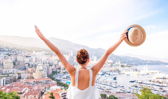 海と街を見渡せる高台で女性が両手を広げて立つ後ろ姿。旅が生き方の選択肢を広げるイメージ